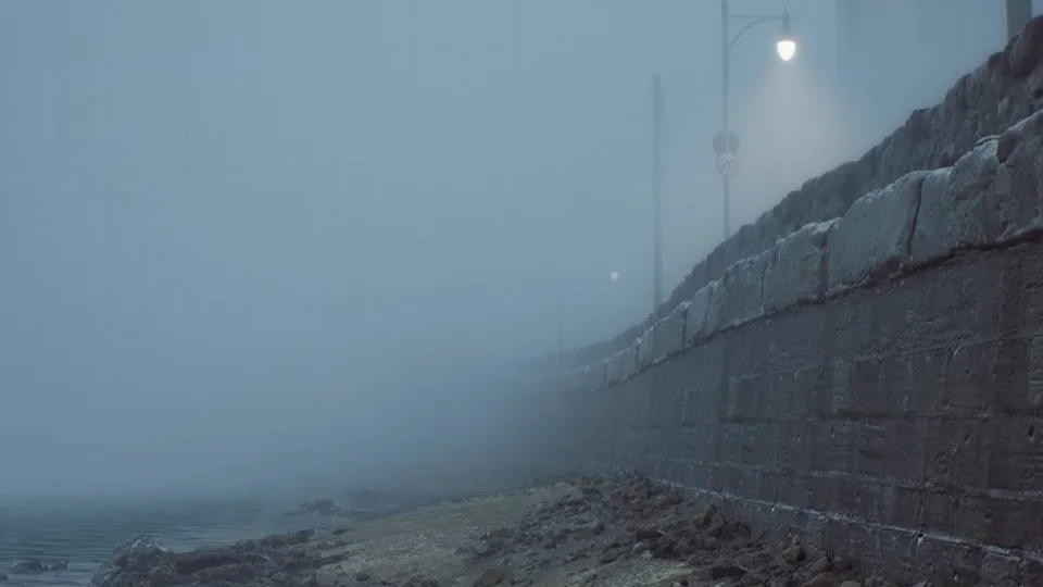 A rocky shoreline beside a stone seawall and a streetlamp in thick fog